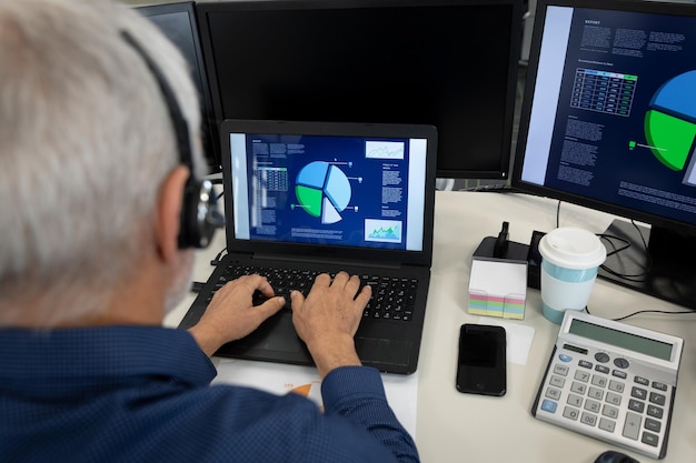 Kaukaski biznesmen pracujący w nowoczesnym biurze, siedzący przy biurku z komputerem, noszący zestaw słuchawkowy do telefonu i korzystający z laptopa. Dystans społeczny w miejscu pracy podczas pandemii Koronawirusa Covid 19.
