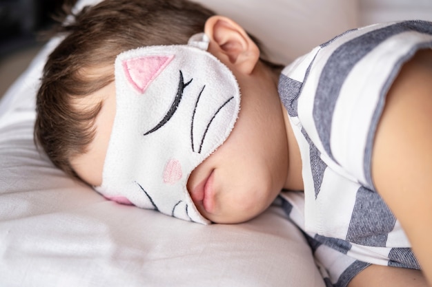 Kaukaski chłopiec w wieku przedszkolnym w piżamie w paski z maską kotka spać w białym łóżku.