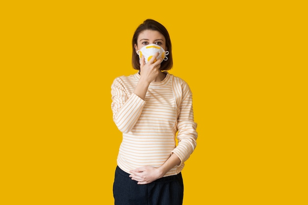 Kaukaski kobieta w ciąży z maską medyczną na twarzy, pozowanie na żółtym tle
