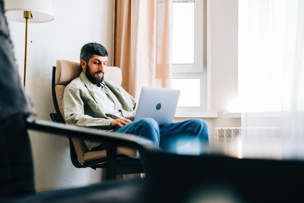 Kaukaski mężczyzna freelancer pracuje w domu, siedząc na fotelu i używając laptopa.