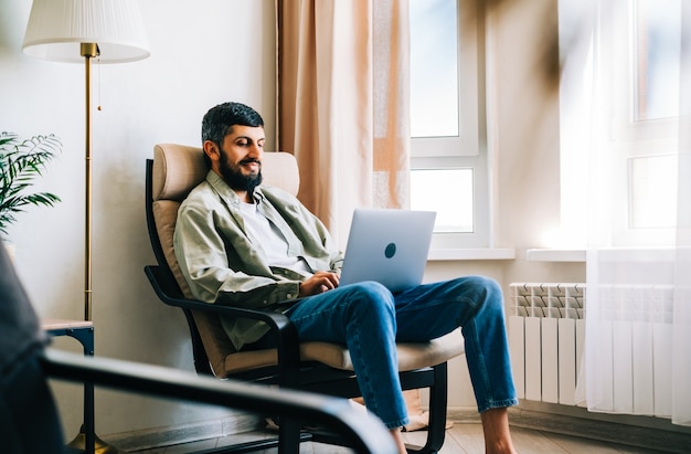 Kaukaski mężczyzna freelancer pracuje w domu, siedząc na fotelu i używając laptopa.