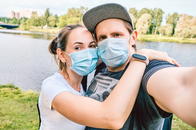 Kaukaski mężczyzna i kobieta z maską medyczną spacerują ulicą w parku podczas pandemii i robią zdjęcie selfie.