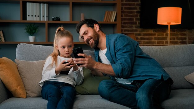 Kaukaski mężczyzna i mała dziewczynka siedzą razem w domu, trzymając nowoczesny telefon, grając w gry online