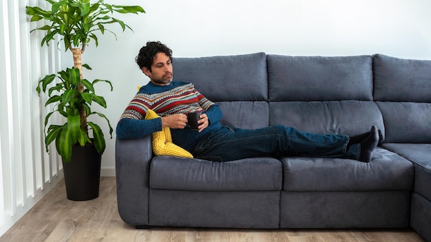 Kaukaski mężczyzna pije poranną kawę expresso, czując się wyczerpany i leżąc na kanapie w domu. Przystojny model mężczyzna czuje się zmęczony po pracy i kawiarni.