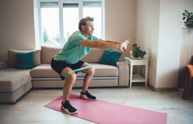 Kaukaski mężczyzna robi fitness w domu za pomocą specjalnych taśm na podłodze podczas kucania