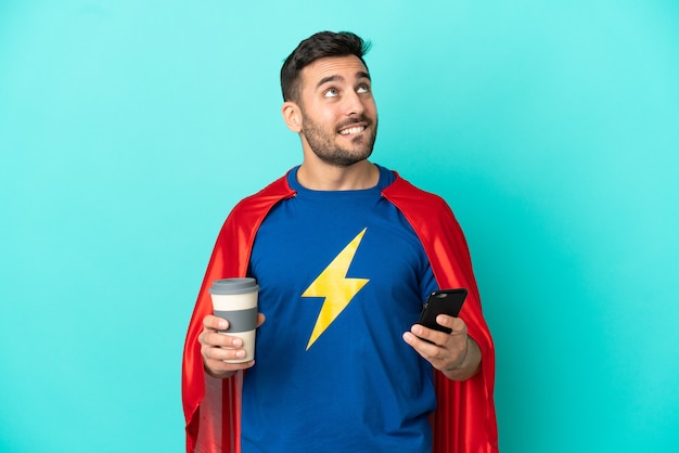 Kaukaski mężczyzna Super Hero na białym tle na niebieskim tle trzymający kawę na wynos i telefon komórkowy, myśląc o czymś