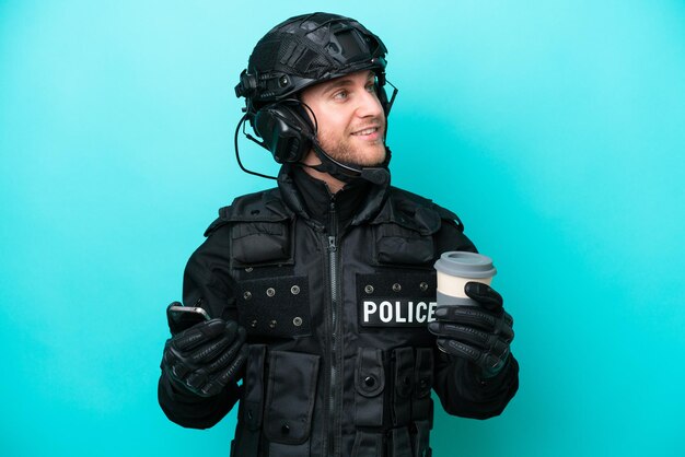 Kaukaski mężczyzna SWAT odizolowany na niebieskim tle trzymający kawę na wynos i telefon komórkowy