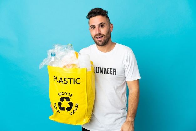 Kaukaski mężczyzna trzyma torbę pełną plastikowych butelek do recyklingu na białym tle na niebiesko z wyrazem twarzy zaskoczenia