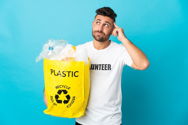 Kaukaski Mężczyzna Trzymający Torbę Pełną Plastikowych Butelek Do Recyklingu Na Białym Tle Na Niebieskim Tle, Mający Wątpliwości I Myślący