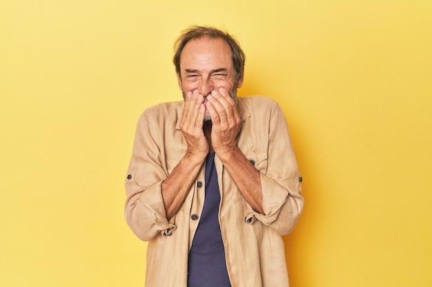 Kaukaski mężczyzna w średnim wieku w żółtym studiu śmiejący się z czegoś zakrywającego usta rękami