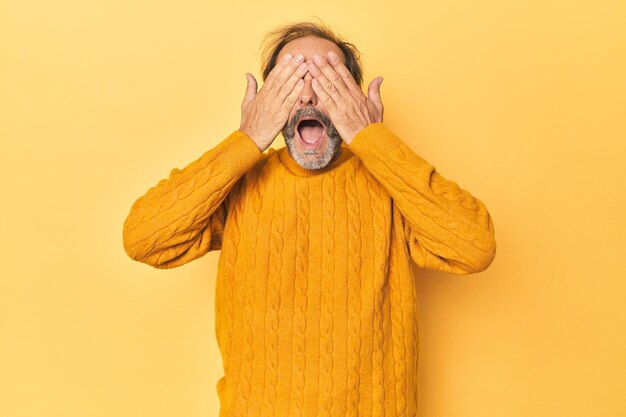 Zdjęcie kaukaski mężczyzna w średnim wieku w żółtym studiu zakrywa oczy rękami i uśmiecha się szeroko, czekając