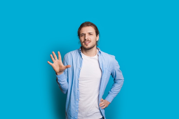 Kaukaski mężczyzna z długimi włosami i brodą, wskazując numer 5 dłonią, pozując na ścianie w niebieskim studio