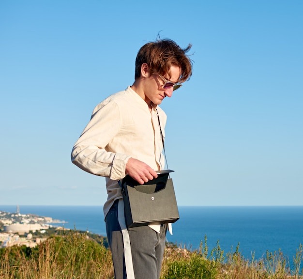 Kaukaski mężczyzna z Hiszpanii, stojący na trawiastym wzgórzu nad oceanem i zamykający czarną torebkę
