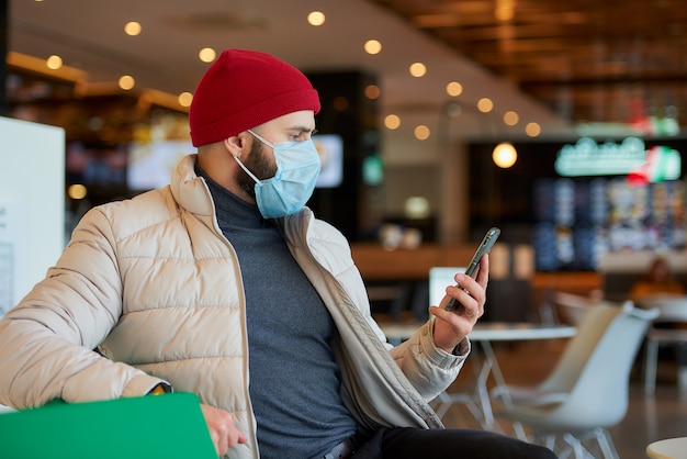 Kaukaski Mężczyzna Z Maską Chirurgiczną Na Twarzy Za Pomocą Smartfona W Centrum Handlowym.