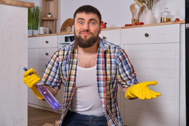 Kaukaski młody brodaty mężczyzna siedzi na podłodze w swojej kuchni i próbuje ją wyczyścić, używając wszystkich swoich ...