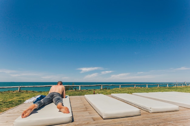 Kaukaski młody człowiek bez koszuli i dżinsach leżąc na plaży na białym materacu