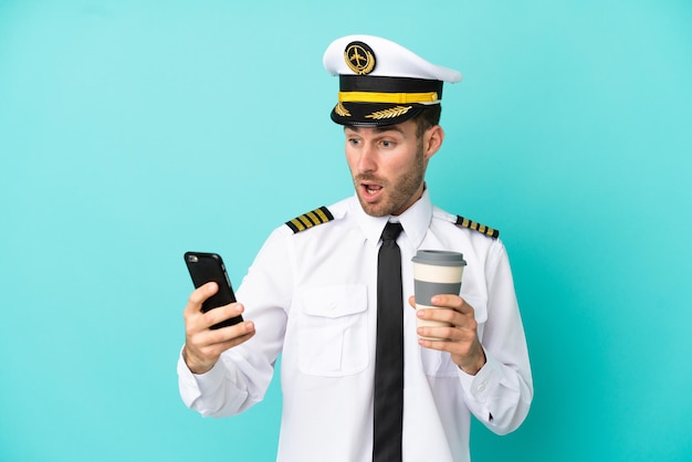 Kaukaski pilot samolotu na białym tle na niebieskim tle trzymający kawę na wynos i telefon komórkowy