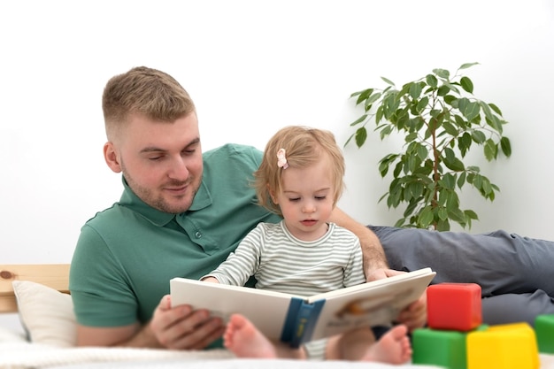 Kaukaski Przystojny Brodaty Mężczyznamała Urocza Dziewczynka Czyta Książkę Na łóżku W Pomieszczeniu Na Białym Tleszczęśliwa Rodzinaojciec I Intant Spędzają Razem Wolny Czaszwiązek Rodzic-dziecko