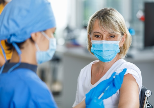 Kaukaski starszy pacjent nosi maskę, patrząc w oczekiwaniu na zastrzyk szczepienia, podczas gdy lekarz w niebieskim mundurze szpitalnym przygotowuje szczepionkę z dawki szklanej fiolki w niewyraźne na pierwszym planie.