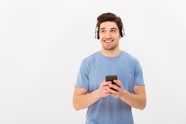 Kaukaski uśmiechnięty mężczyzna w przypadkowej koszulce słucha muzyki przez bezprzewodowe słuchawki używać smartphone, odizolowywającego nad biel ścianą