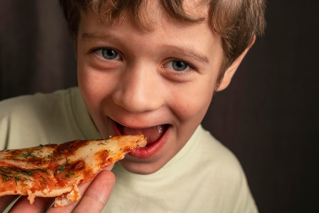 Kaukaski wesoły chłopiec zjada kawałek pizzy i patrzy w kamerę z uśmiechem