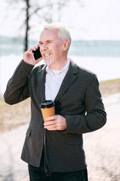 Kawa w parku. Optymistycznie starszy biznesmen pije kawę w parku i rozmawia przez telefon