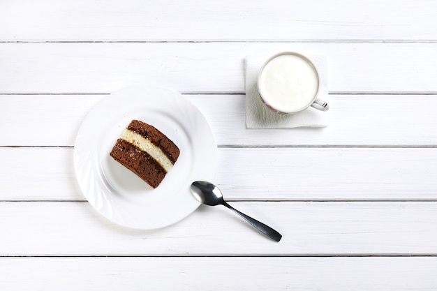 Kawałek brązowego ciasta na łyżce talerzowej i filiżance z kefirem jako śniadanie na drewnianej białej powierzchni