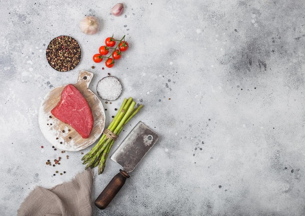 Zdjęcie kawałek surowej wołowiny stek sirlion na okrągłej desce do krojenia z pomidorami, czosnkiem i szparagami oraz siekierą na jasnym stole w kuchni.