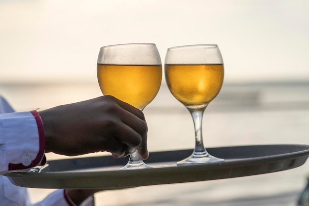 Kelner podaje dwa kieliszki białego wina na tacy w restauracji na plaży podczas zachodu słońca z bliska