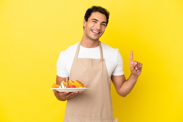 Kelner w restauracji trzymający gofry na izolowanym żółtym tle pokazujący i unoszący palec na znak najlepszych