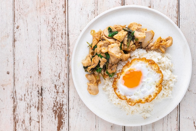 Khao Pad Ka Prao Gai Kai Dao, tajskie jedzenie, przesyłany strumieniowo ryż z bazyliowym smażonym kurczakiem i jajkiem sadzonym w białej ceramicznej płycie na białym starym tle tekstury drewna z kopią miejsca, widok z góry