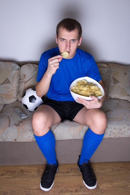 Kibic w mundurze oglądający piłkę nożną w telewizji w domu i jedzący chipsy