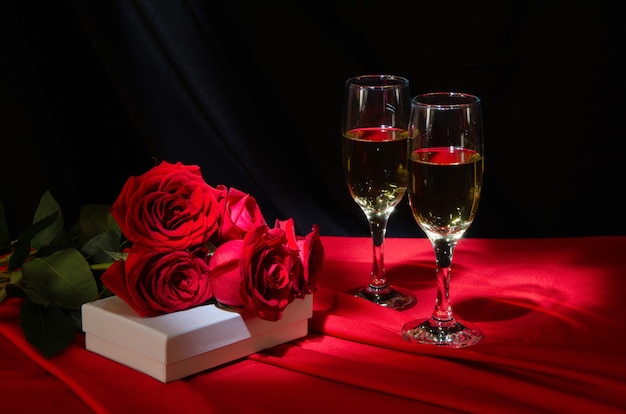 Kieliszki z szampanem, czerwonymi różami i prezentem na czerwonym obrusie.