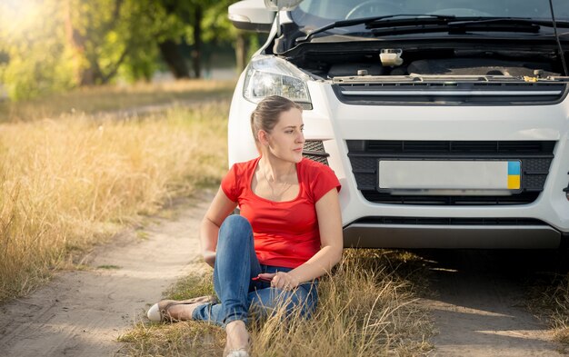 Zdjęcie kierowca siedząca na ziemi obok zepsutego samochodu i wzywająca pomocy