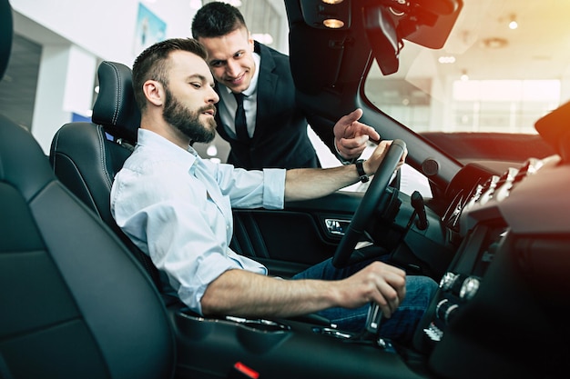 Kierownik dealera w garniturze z uśmiechem pokazuje młodemu kupcowi wszystkie możliwości techniczne samochodu