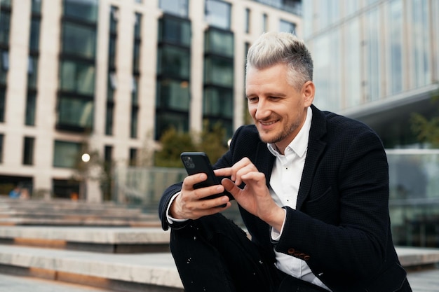 Kierownik siedzi w pobliżu centrum biznesowego i pisze wiadomość przez telefon