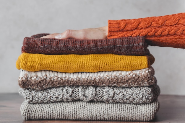 Kilka dzianinowych, ciepłych swetrów, kobieca dłoń na stosie ubrań w jesiennej palecie kolorów.