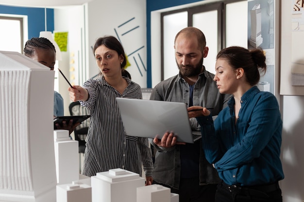 Kilku profesjonalnych architektów trzymających laptopa patrzących na plany obok kolegów z tabletem wskazującym na makietę 3d wieżowca. Zespół inżynierów architektonicznych wykonujących twórczą pracę zespołową.
