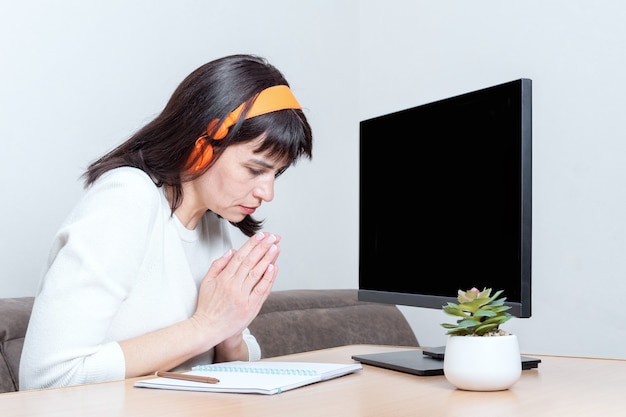 Kłaniająca się kobieta w białym swetrze i słuchawkach siedząca przy stole patrzy na monitor z pustym czarnym ekranem i składa dłonie w geście podziękowania, wdzięczności