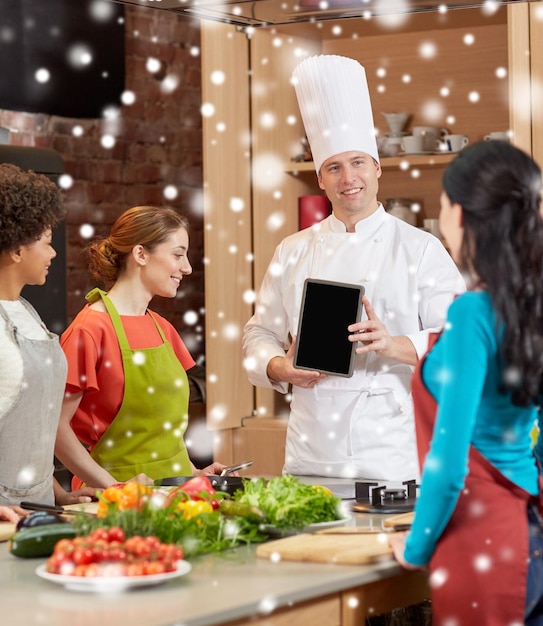 Zdjęcie klasa gotowania, koncepcja kulinarna, jedzenie, technologia i ludzie - szczęśliwe kobiety z kucharzem szefem kuchni pokazujące pusty ekran komputera typu tablet w kuchni nad efektem śniegu