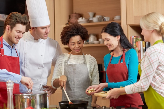 klasa gotowania, koncepcja kulinarna, żywnościowa i ludzie - szczęśliwa grupa przyjaciół i kucharz kucharz gotujący w kuchni