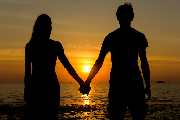 Klasyczna scena walentynkowa z sylwetką młodej pary trzymającej się za ręce podczas kontemplacji zachodu słońca na plaży.