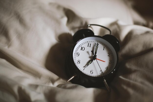 Klasyczny czarny budzik pokazujący godzinę siódmą z łóżkiem w tle