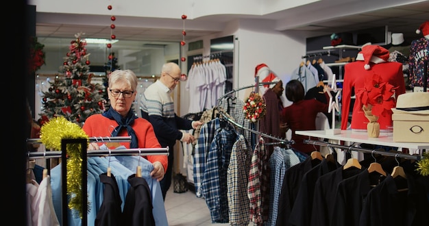 Klienci wędrują po zajętym sklepie z odzieżą ozdobionym świątecznymi ozdobami podczas zimowych świątecznych uroczystości. Klienci kupują prezenty podczas promocyjnych sprzedaży xmas w butiku mody