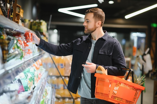 Klient w supermarkecie mężczyzna robi zakupy spożywcze stojąc z koszykiem wybierając produkt spożywczy w pomieszczeniu facet kupujący artykuły spożywcze w sklepie spożywczym selektywna ostrość kopia przestrzeń