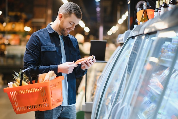 Klient w supermarkecie mężczyzna robi zakupy spożywcze stojąc z koszykiem wybierając produkt spożywczy w pomieszczeniu facet kupujący artykuły spożywcze w sklepie spożywczym selektywna ostrość kopia przestrzeń