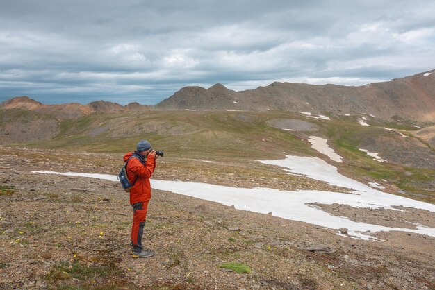 Klimatyczna sceneria z mężczyzną w czerwieni na tle pasma górskiego z ostrymi skałami pod szarym pochmurnym niebem Turysta z aparatem fotografuje dramatyczny górski krajobraz Mężczyzna fotografuje góry przy ponurej pogodzie