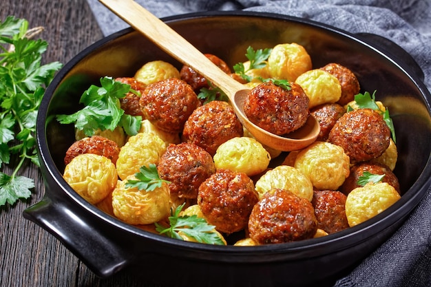 Klopsiki i kulki spaghetti zapiekane na czarnym naczyniu do pieczenia - kuchnia włoska, podawane na ciemnym drewnianym tle ze świeżą pietruszką i drewnianą łyżką, widok z góry, zbliżenie