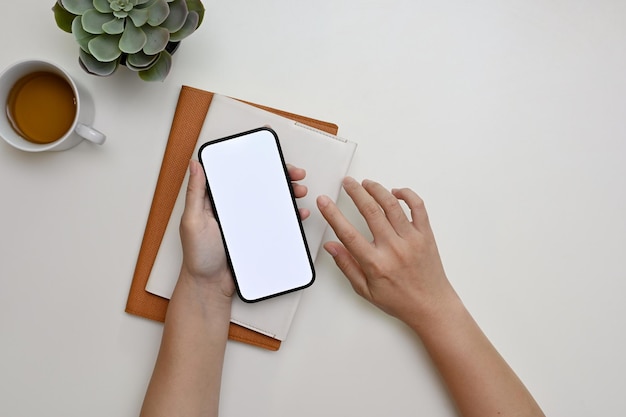 Zdjęcie kobieca dłoń trzymająca makietę smartfona nad minimalnym białym widokiem z góry biurka