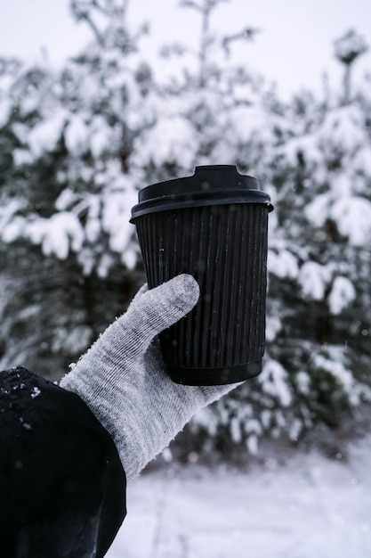 Kobieca dłoń w zimowych ciepłych rękawiczkach trzymająca czarny eko papierowy kubek Zimowy piknik na świeżym powietrzu Kreatywny modny kubek recyklingu zero odpadów Makieta Gorący napój rozgrzewający na wynos w zimowy dzień Koncepcja na wynos
